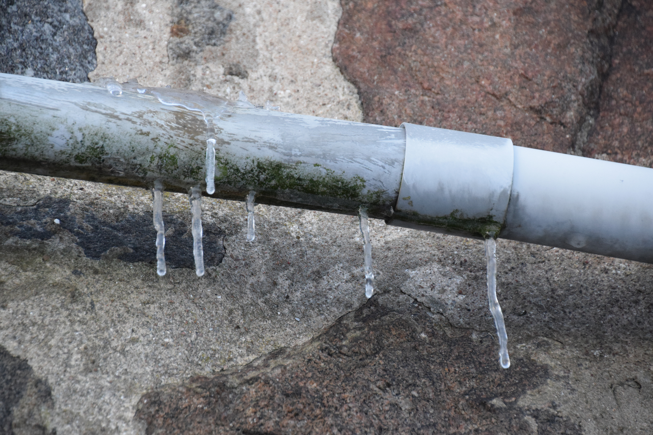 A frozen pipe with hanging icicles against a granite stone wall